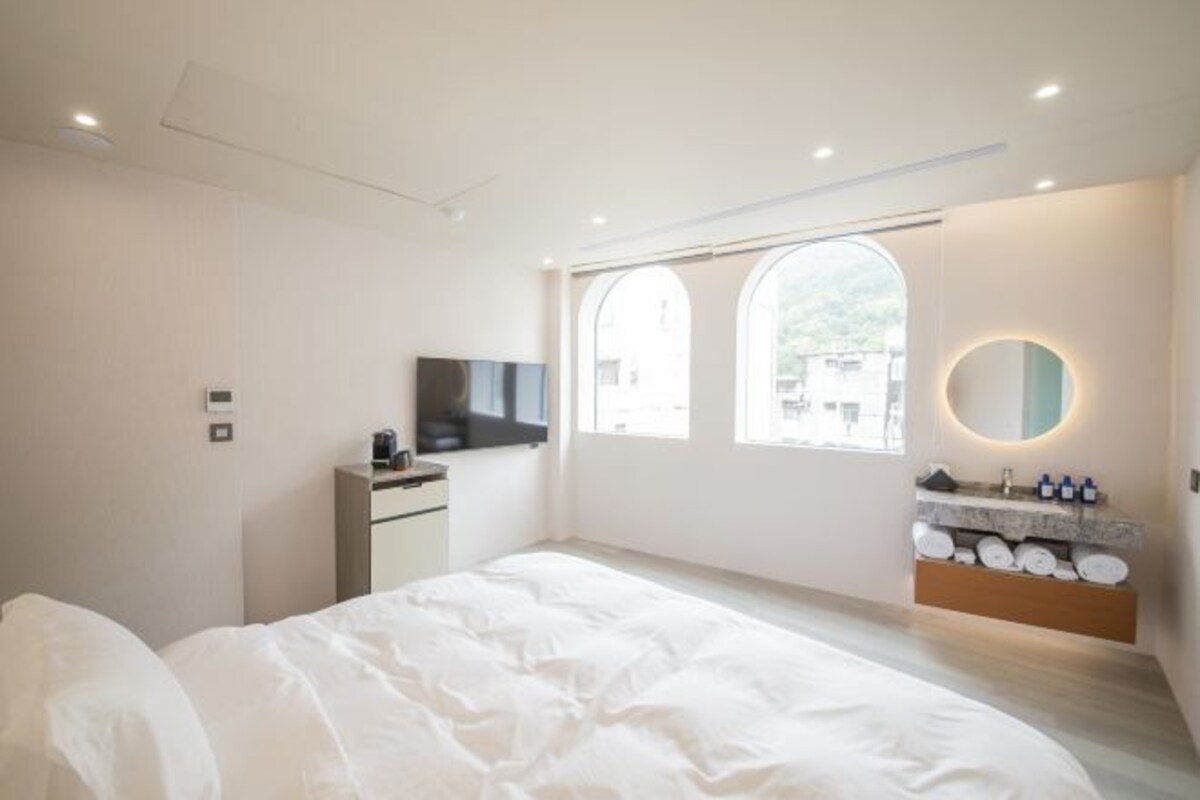 A modern hotel room with a large bed, a TV, a vanity with a mirror, and a window with a view of the outside.