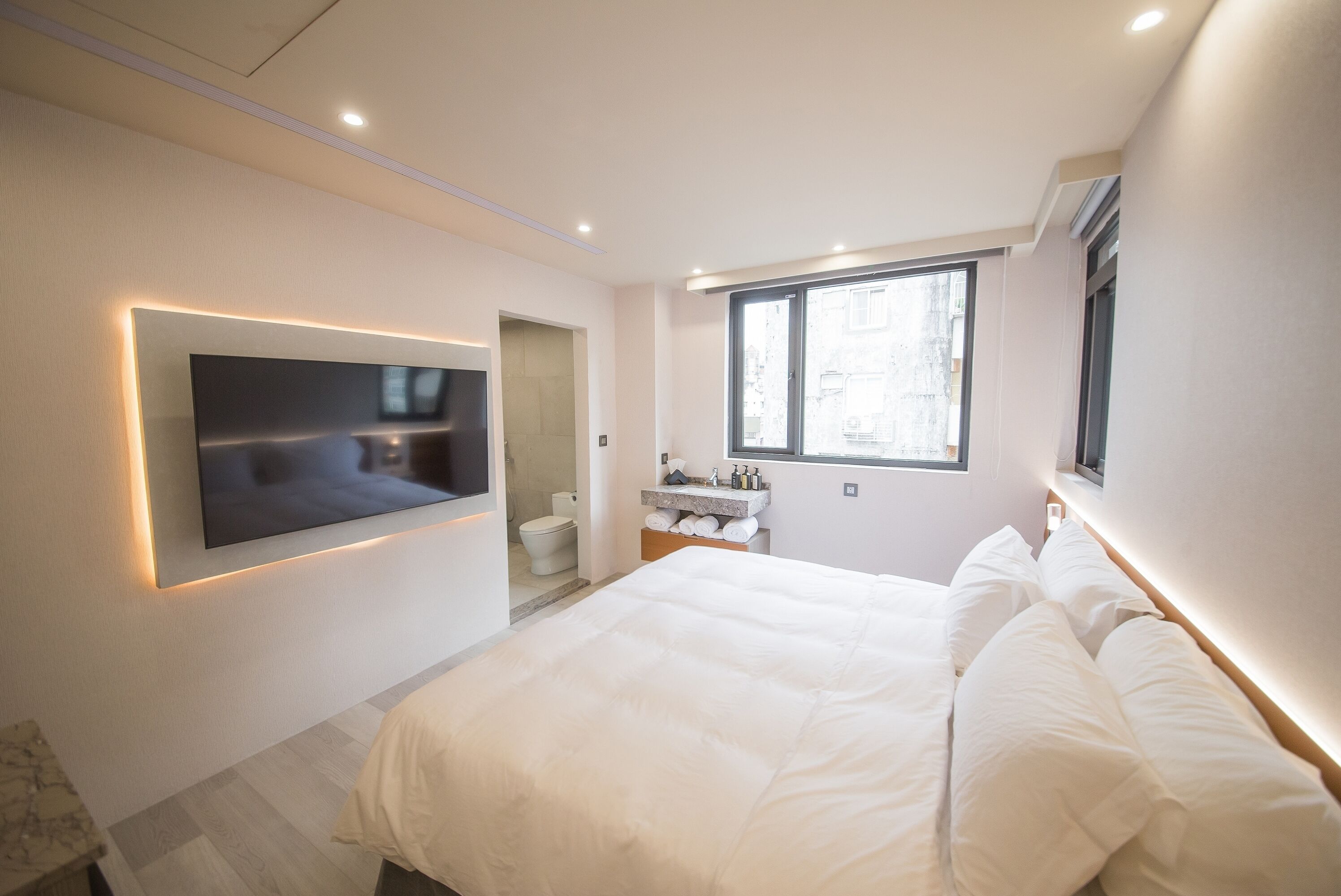 Un dormitorio moderno con una cama grande, un televisor de pantalla plana en la pared, una ventana y un baño pequeño.