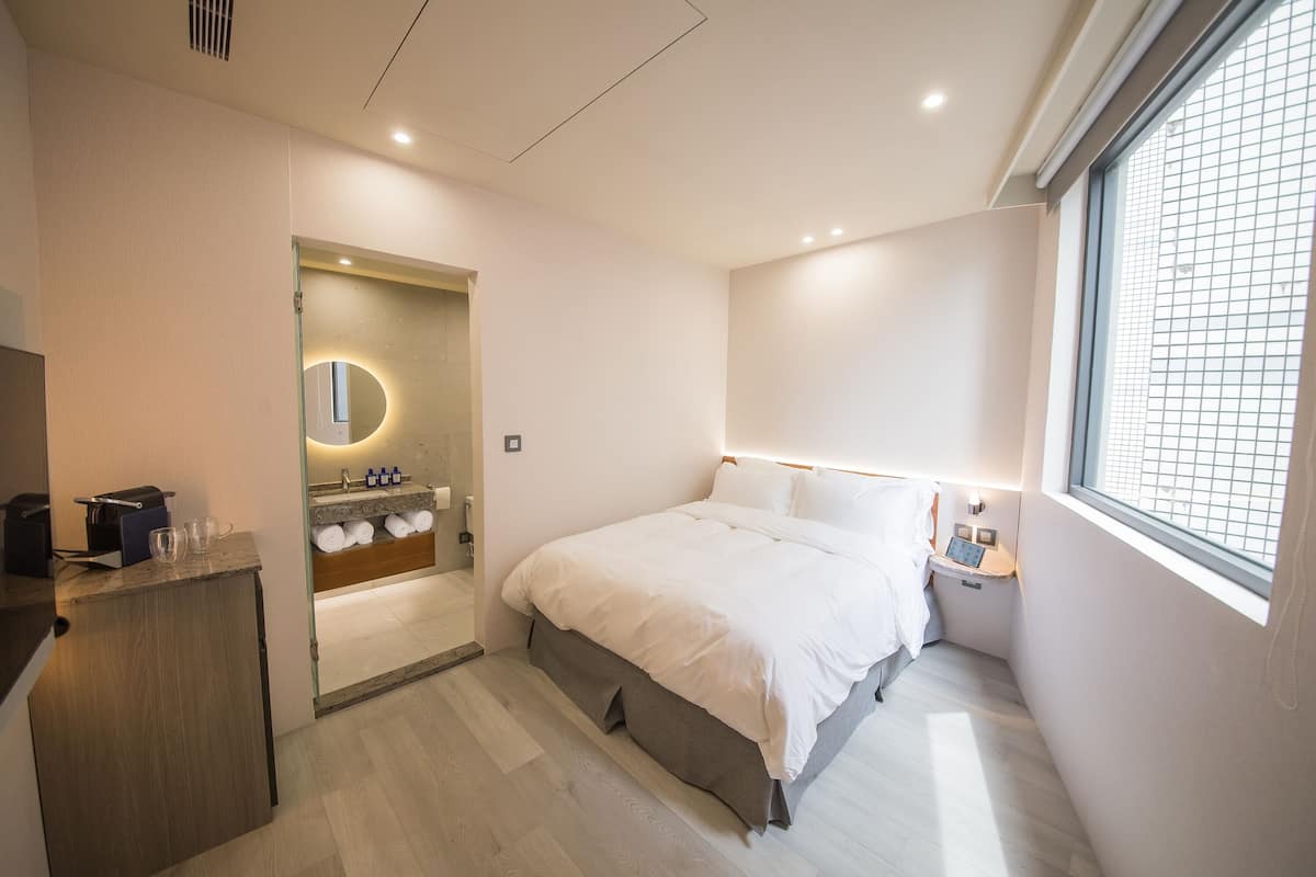 Habitación de hotel moderna con una cama grande, una mesita de noche con lámpara, un espejo integrado y una ventana con vistas a la ciudad.