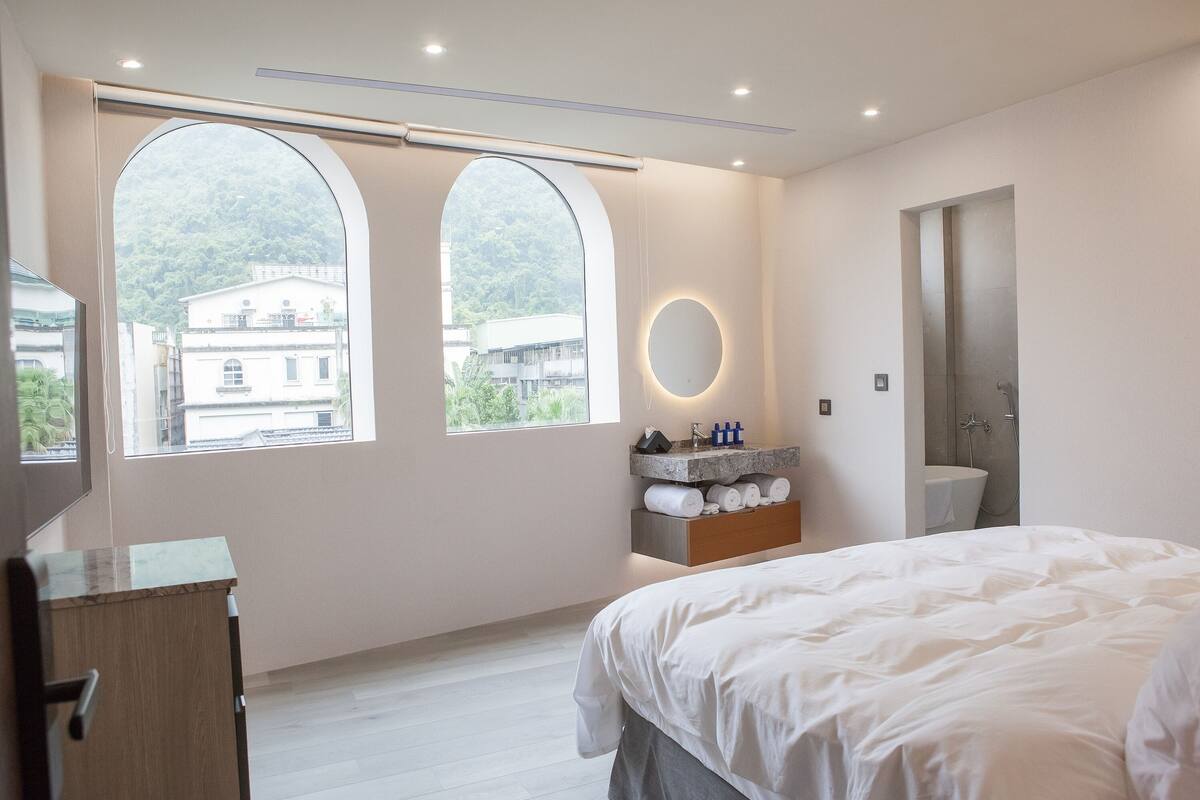 Una habitación de hotel moderna con una cama grande, una tocador con espejo y vista a un edificio a través de ventanales arqueados.
