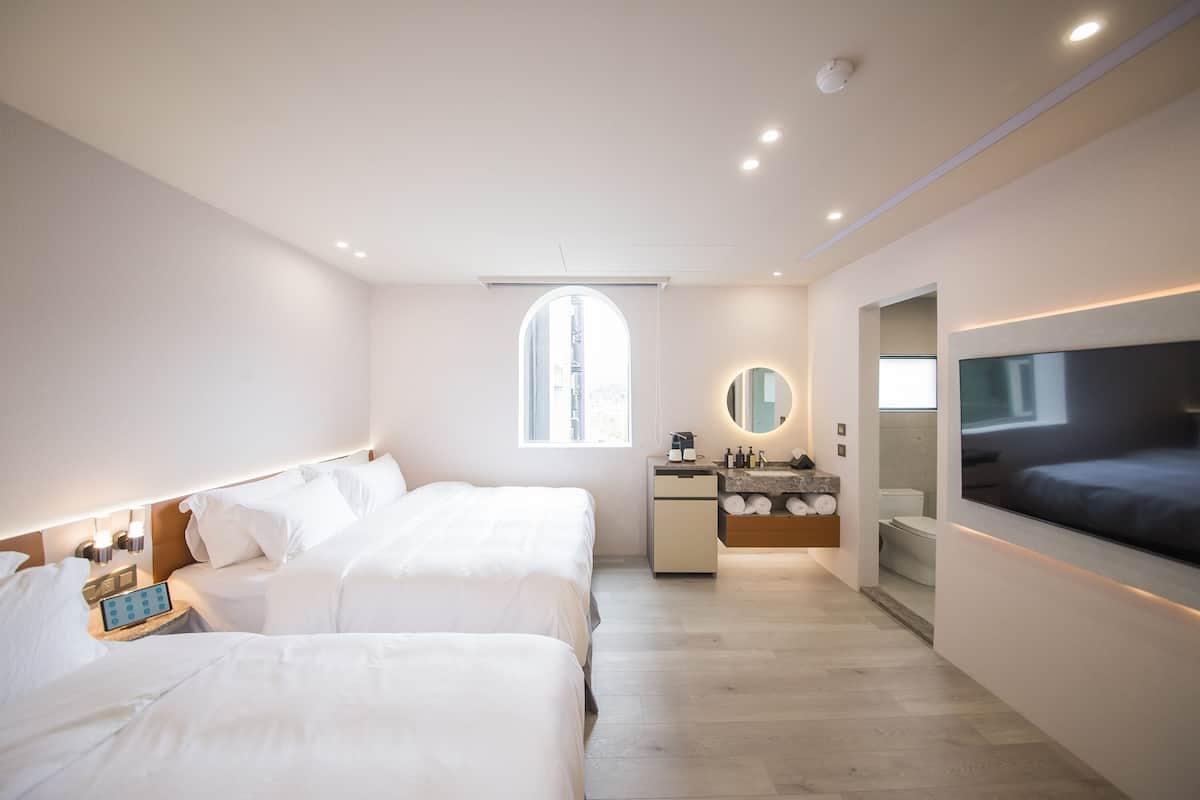 Una habitación de hotel moderna con dos camas, un baño, un televisor y un ventanal amplio.
