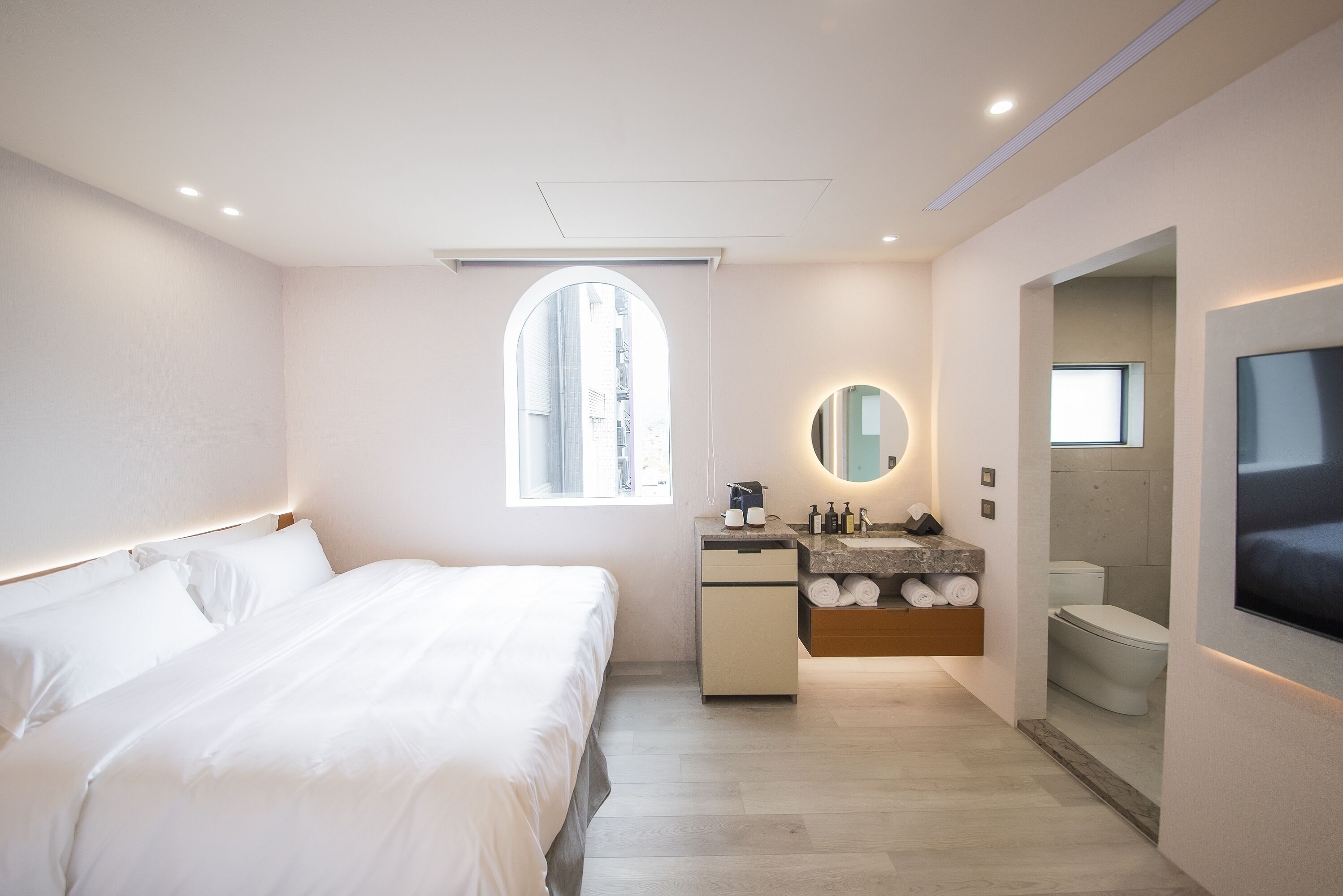 A modern hotel room with a large bed, a vanity with a mirror, a television, and a window with a view of buildings.