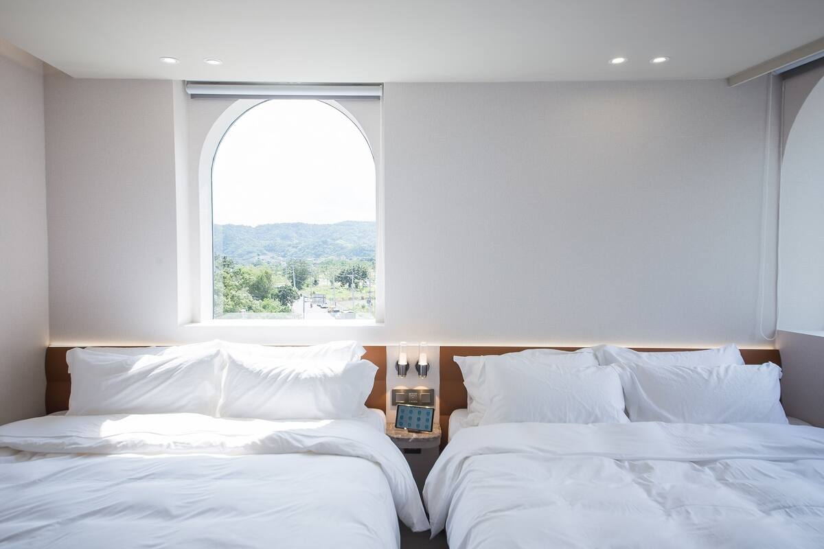 Una habitación de hotel con dos camas, un ventanal arqueado grande y una mesita de noche con un reloj.
