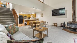 Uma sala de estar ampla com sofá, mesa de centro de madeira, lareira, cozinha ao fundo e escada que leva a um andar superior.