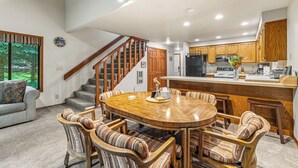 Uma sala de estar ampla com uma mesa de jantar de madeira cercada por cadeiras listradas, uma escada com corrimão de madeira e uma cozinha com armários de madeira e uma geladeira preta.