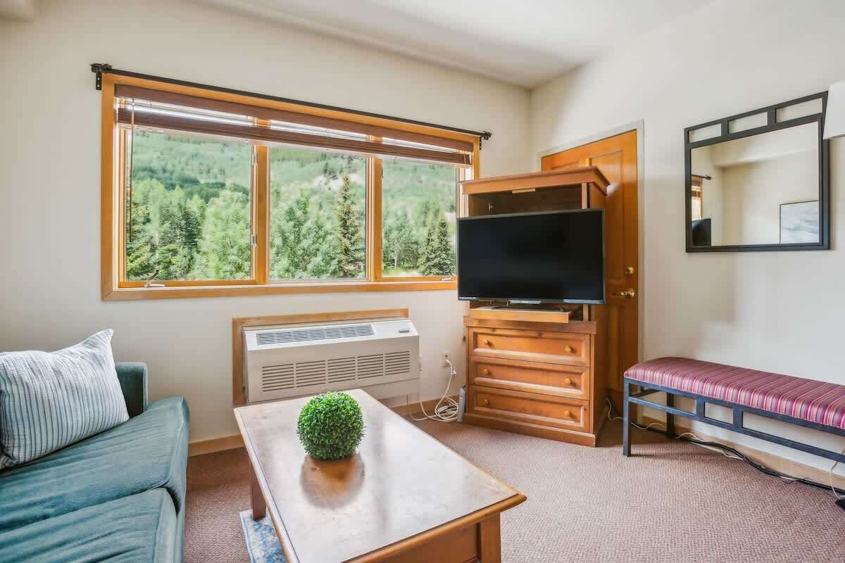 Ein Zimmer mit großem Fenster, Fernseher, Holzschrank, einer Bank und einem kleinen Tisch mit Pflanze.