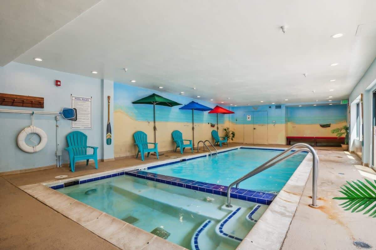 An indoor swimming pool with blue chairs and umbrellas, a lifebuoy, and a pool rules sign.