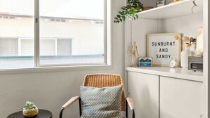 A room with a wicker chair, a small black table, a woven rug, a white cabinet with shelves, and a window with blinds. - South Mission Beach Bungalow (San Diego)