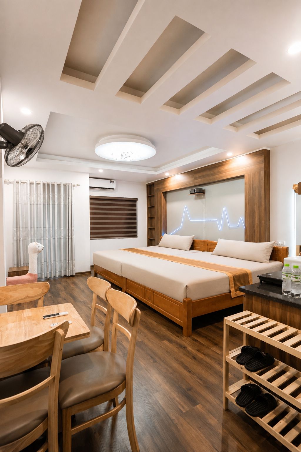 A modern hotel room with a wooden bed, a desk, chairs, and a shoe rack.
