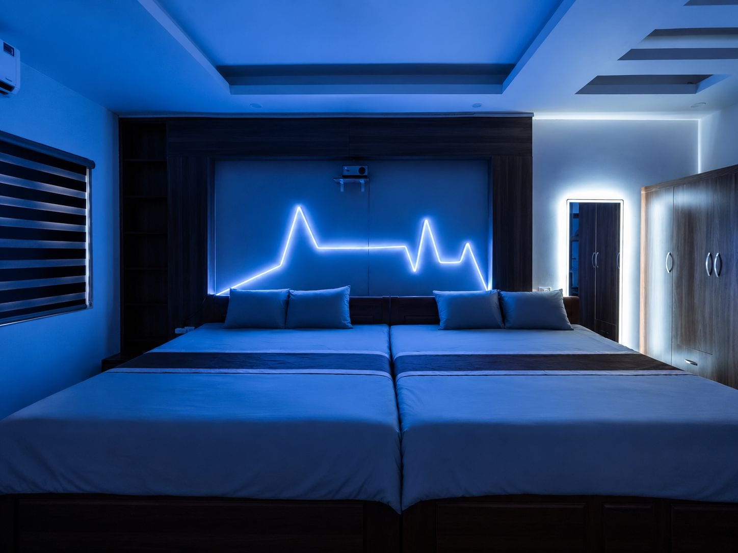 A bedroom with two beds, a neon light wall art, and a wardrobe.