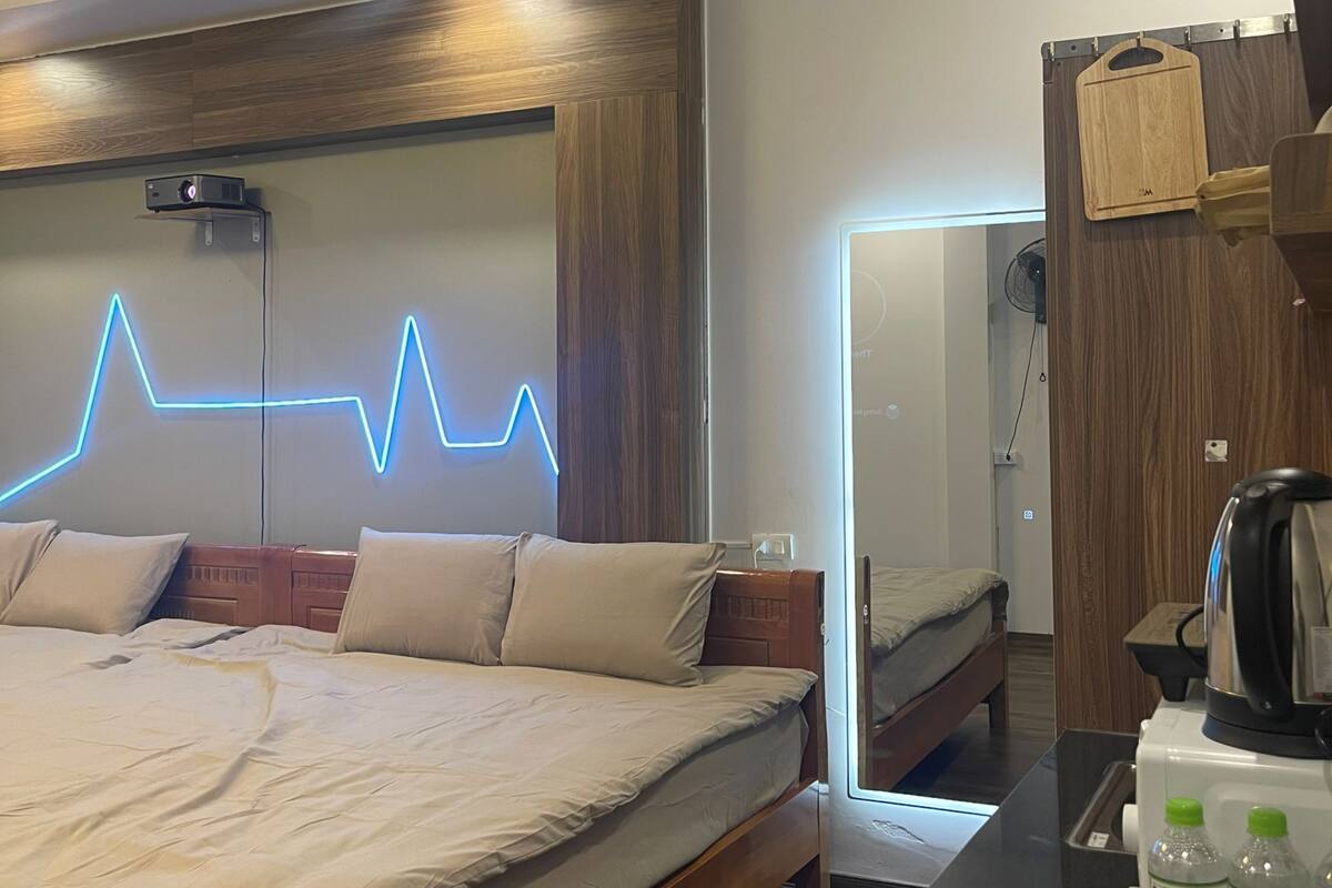 A modern hotel room with a large bed, a wooden headboard, a neon light wall design, a wooden wardrobe, and a small desk with a kettle and water bottles.