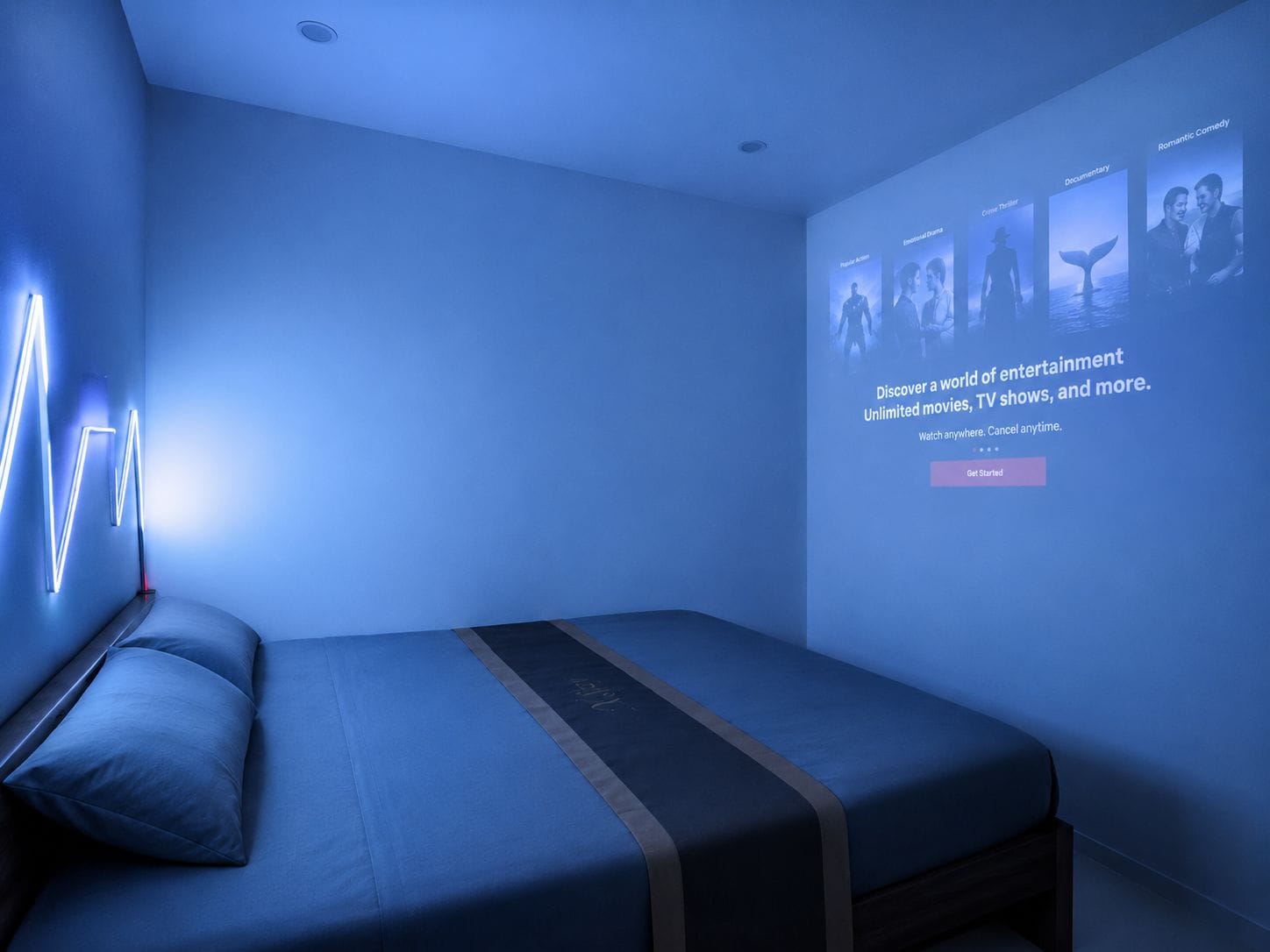 A modern hotel room with a bed, a wall-mounted projector displaying movie genres, and a neon light design on the wall.