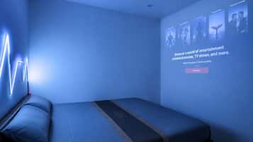 A modern hotel room with a bed, a wall-mounted projector displaying movie genres, and a neon light design on the wall.