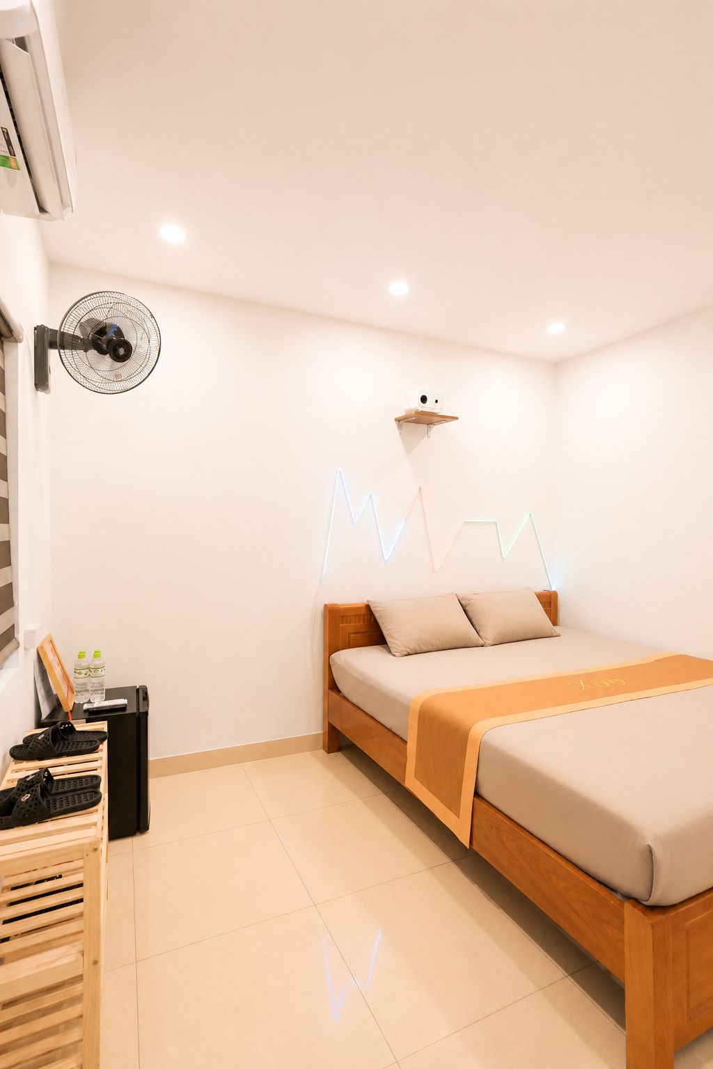 Un dormitorio moderno con una cama, un ventilador de pared, un estante con una cámara y un zapatero.