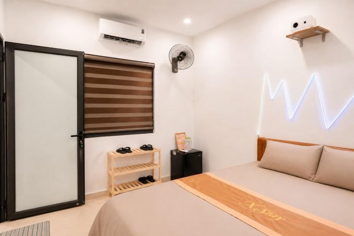Ein Schlafzimmer mit einem Bett, einer Schiebetür, einem Schuhregal, einem Wandventilator und einer Neonleuchte.