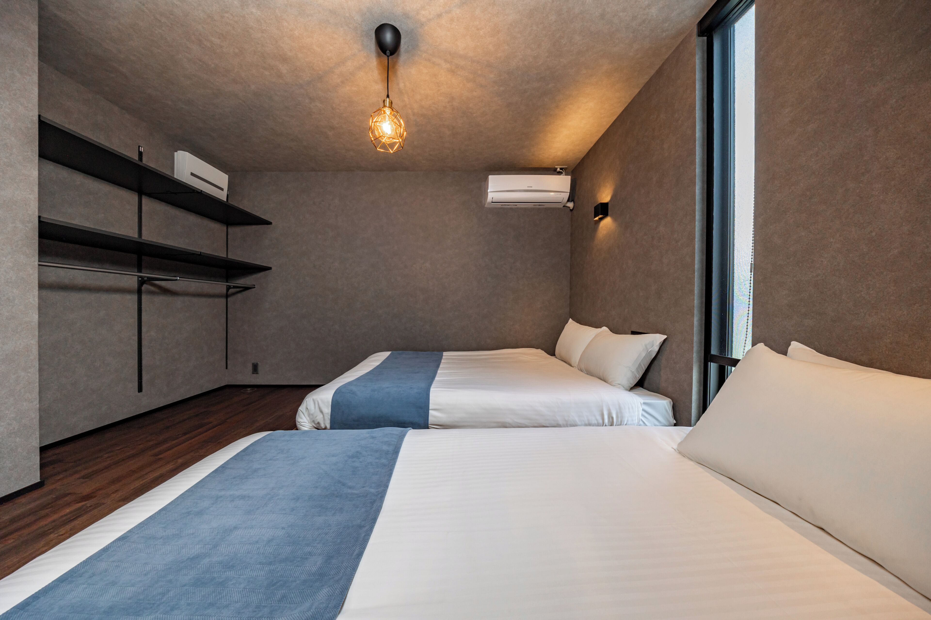 Una habitación de hotel moderna con dos camas, una lámpara de techo, estanterías integradas y un equipo de aire acondicionado.