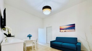 A modern living room with a teal sofa, a white dining table with chairs, a white coffee table, and a painting of a sunset over the ocean.