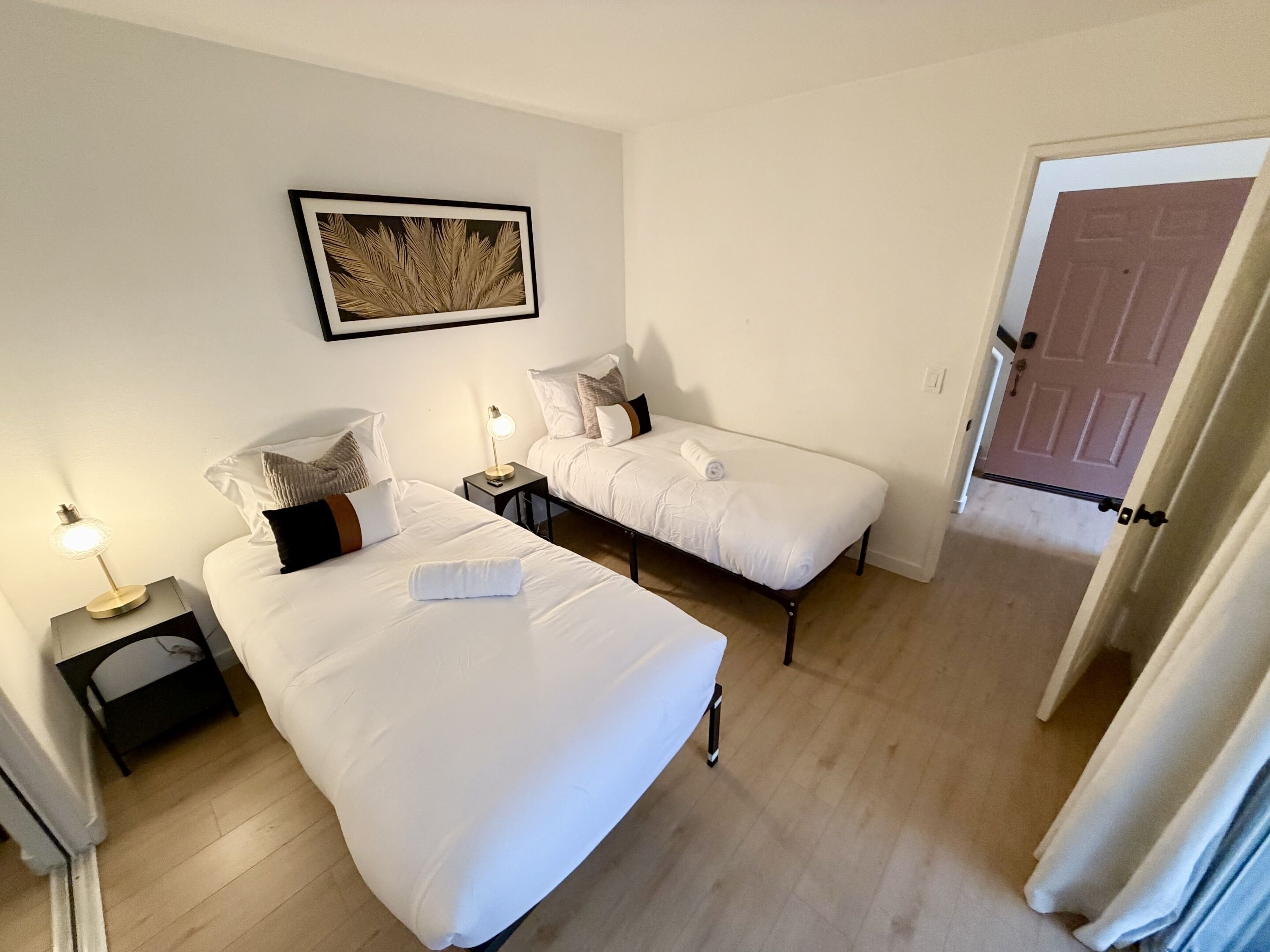 A hotel room with two beds, a bedside table with a lamp, a framed picture on the wall, and a door leading to another room.