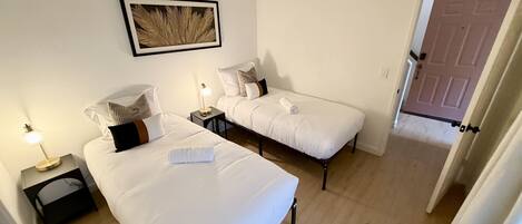 A hotel room with two beds, a bedside table with a lamp, a framed picture on the wall, and a door leading to another room.