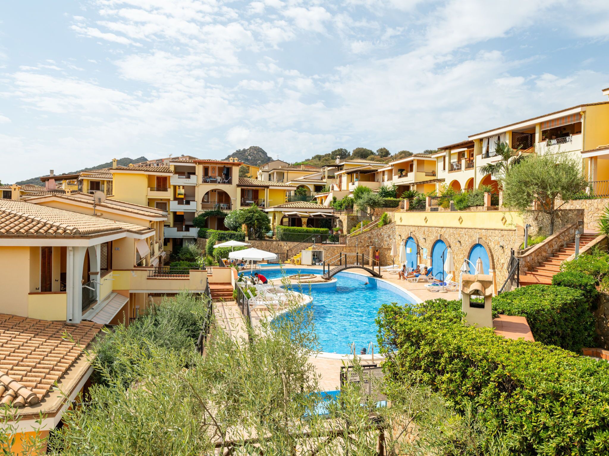 Un resort con piscina central rodeada de edificios de varios pisos con balcones, una escalera de piedra y abundante vegetación.