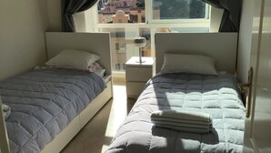 A bedroom with two beds, a window with curtains, a bedside table, and a view of a hillside.