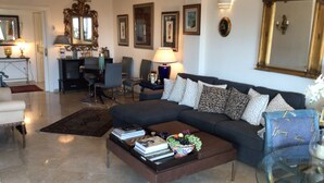 A living room with a dark-colored sofa, a glass coffee table with books and fruit, a dining area with chairs, and a large mirror on the wall.