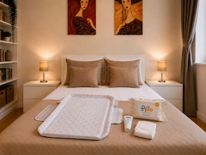 A hotel room with a bed, two bedside lamps, a bedside table, a bookshelf, and two paintings on the wall.