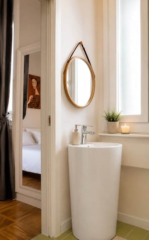 A bathroom with a round mirror, a white sink, a small potted plant, and a candle on a shelf.