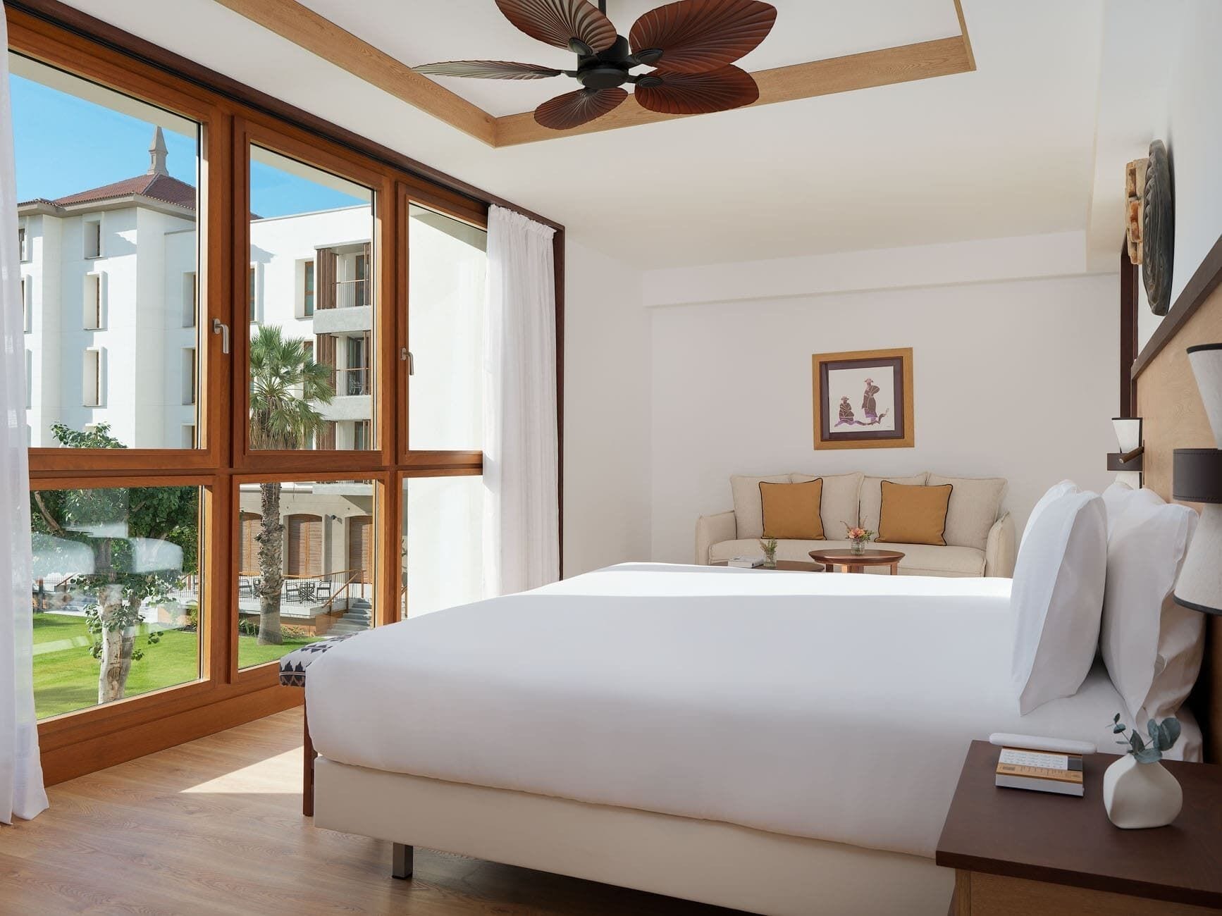 Un dormitorio con una cama grande, un ventilador de techo, un sofá y vistas a un edificio y vegetación exterior.