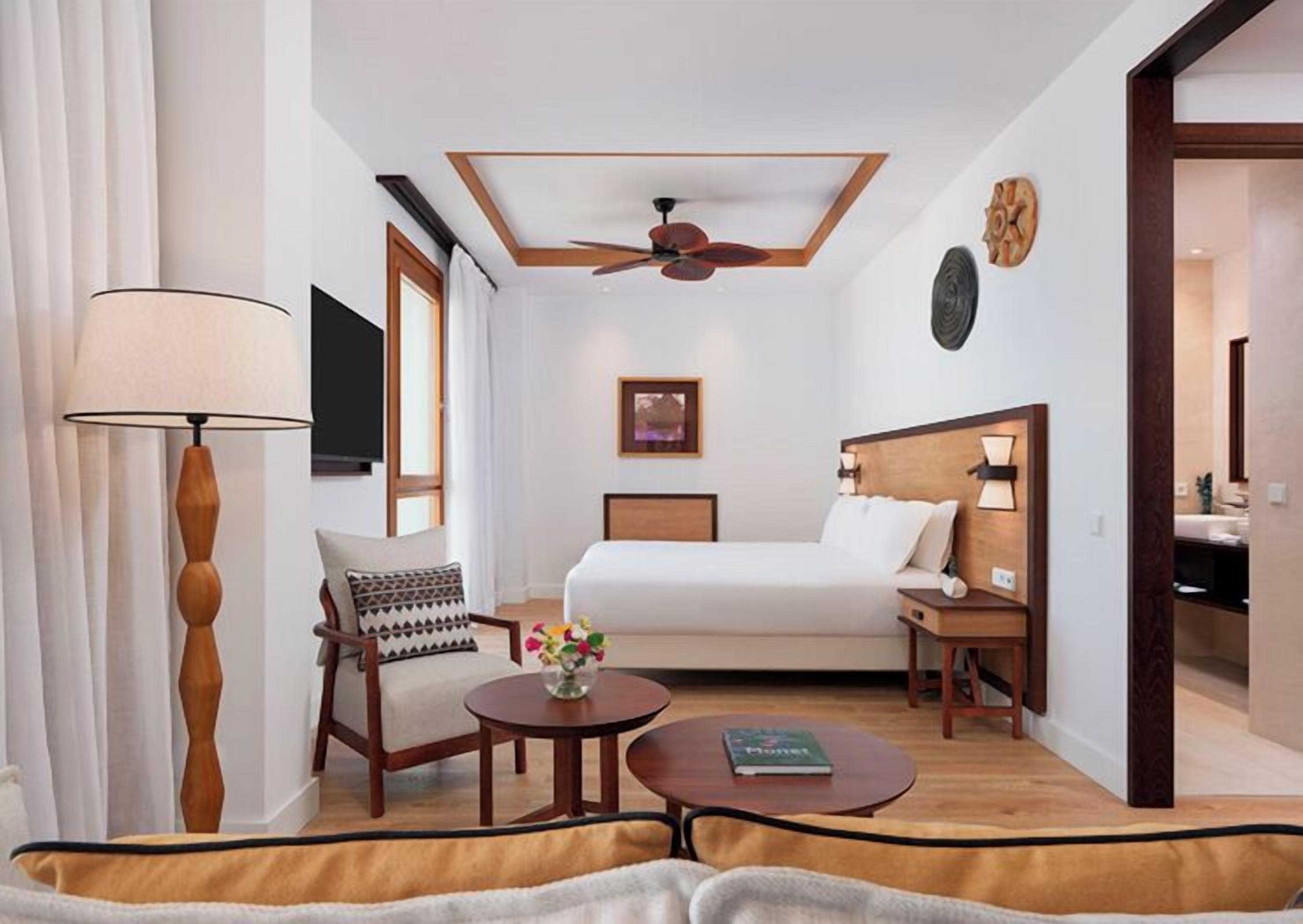 A hotel room with a bed, a chair, a table, a lamp, a ceiling fan, a TV, and a painting on the wall.