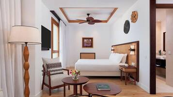 A hotel room with a bed, a chair, a table, a lamp, a ceiling fan, a TV, and a painting on the wall.