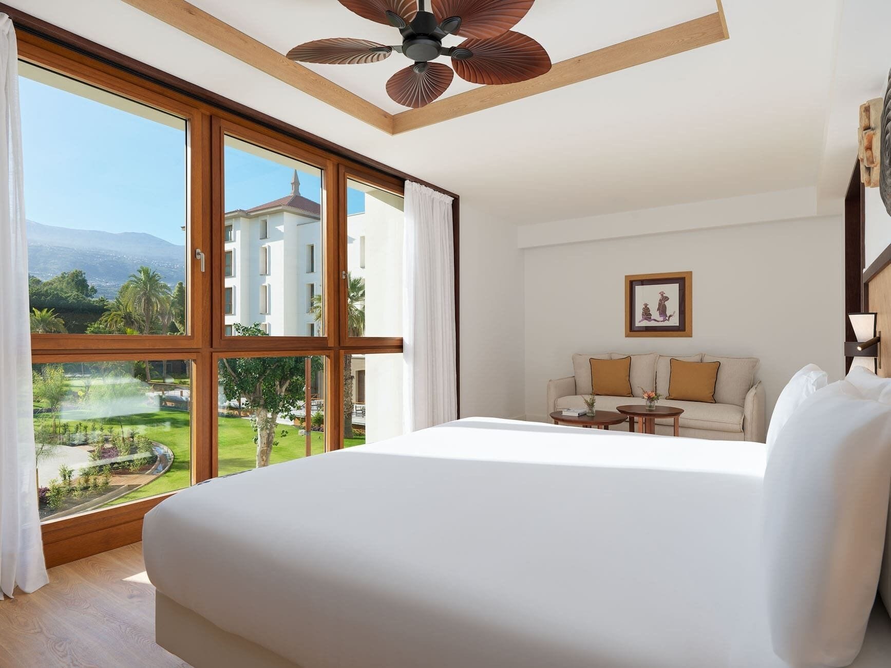A spacious bedroom with a large bed, a ceiling fan, a sofa, and a view of a building and greenery.