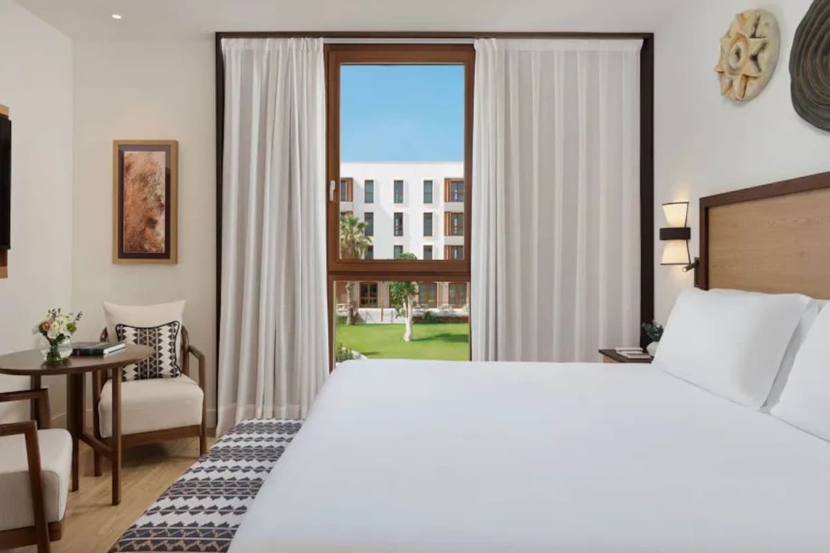 A hotel room with a large bed, a desk with a chair, a window with curtains, and a view of a building outside.