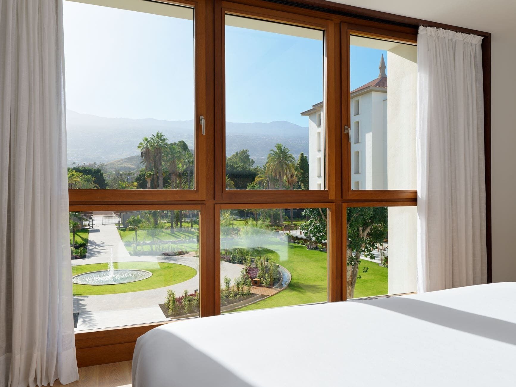 A hotel room with a large window, a bed, and a view of a landscaped garden and distant mountains.