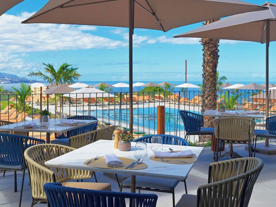 Un área de comedor al aire libre con mesas y sillas, sombrillas y vistas a una piscina y palmeras.