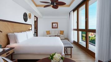 Habitación de hotel con una cama grande, un sofá, una mesita con un florero y un ventilador de techo, además de un ventanal con vistas.
