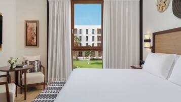 Un cuarto de hotel con una cama grande, un escritorio con una silla, vista de un edificio a través de la ventana y un cuadro en la pared.