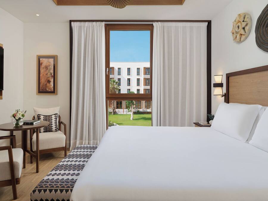 Un cuarto de hotel con una cama grande, un escritorio con una silla, vista de un edificio a través de la ventana y un cuadro en la pared.