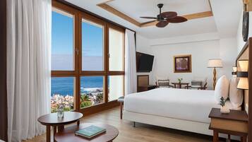 A hotel room with a large window, a bed, a desk, a chair, a ceiling fan, and a TV.