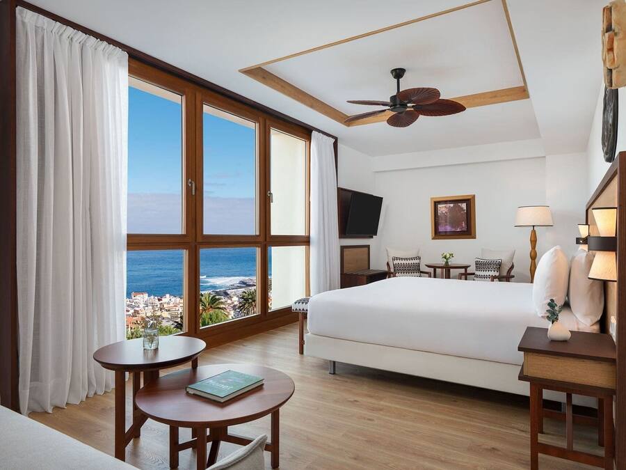 A hotel room with a large window, a bed, a desk, a chair, a ceiling fan, and a TV.