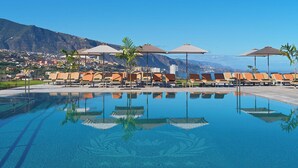 Zona de piscina con sillones reclinables y sombrillas, una piscina cristalina que refleja el entorno y un paisaje montañoso de fondo.