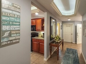 A hallway with a kitchen area, wooden cabinets, a dining table with a vase, and a wall with motivational phrases.