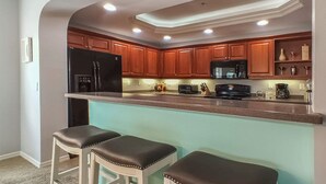 Una cocina con armarios de madera, un refrigerador negro, un microondas, un tostador y una cafetera. Hay dos taburetes de bar con asientos de cuero.