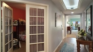 A hallway with a glass door leading to a room with a bed, a blue rug, and a table with a vase. - Gorgeous 3B/2B Island Condo At Bella Lago, Pool, Walk To Beach (Fort Myers Beach)
