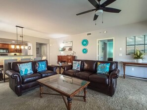 Ein Wohnzimmer mit einem Glastisch, zwei Ledersofas mit blauen Kissen, einem Deckenventilator und einer Küche im Hintergrund.