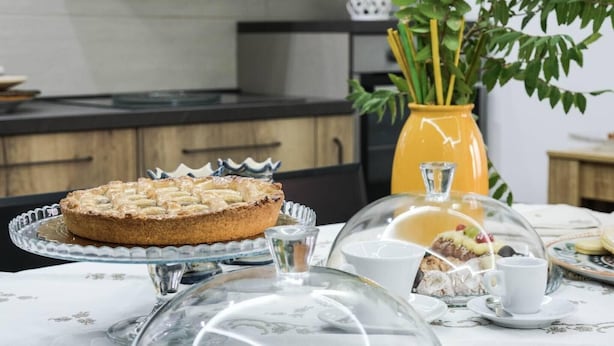 Een ontbijttafel met een taart op een glazen plateau, een gele vaas met planten en een nagerecht onder een glazen stolp.