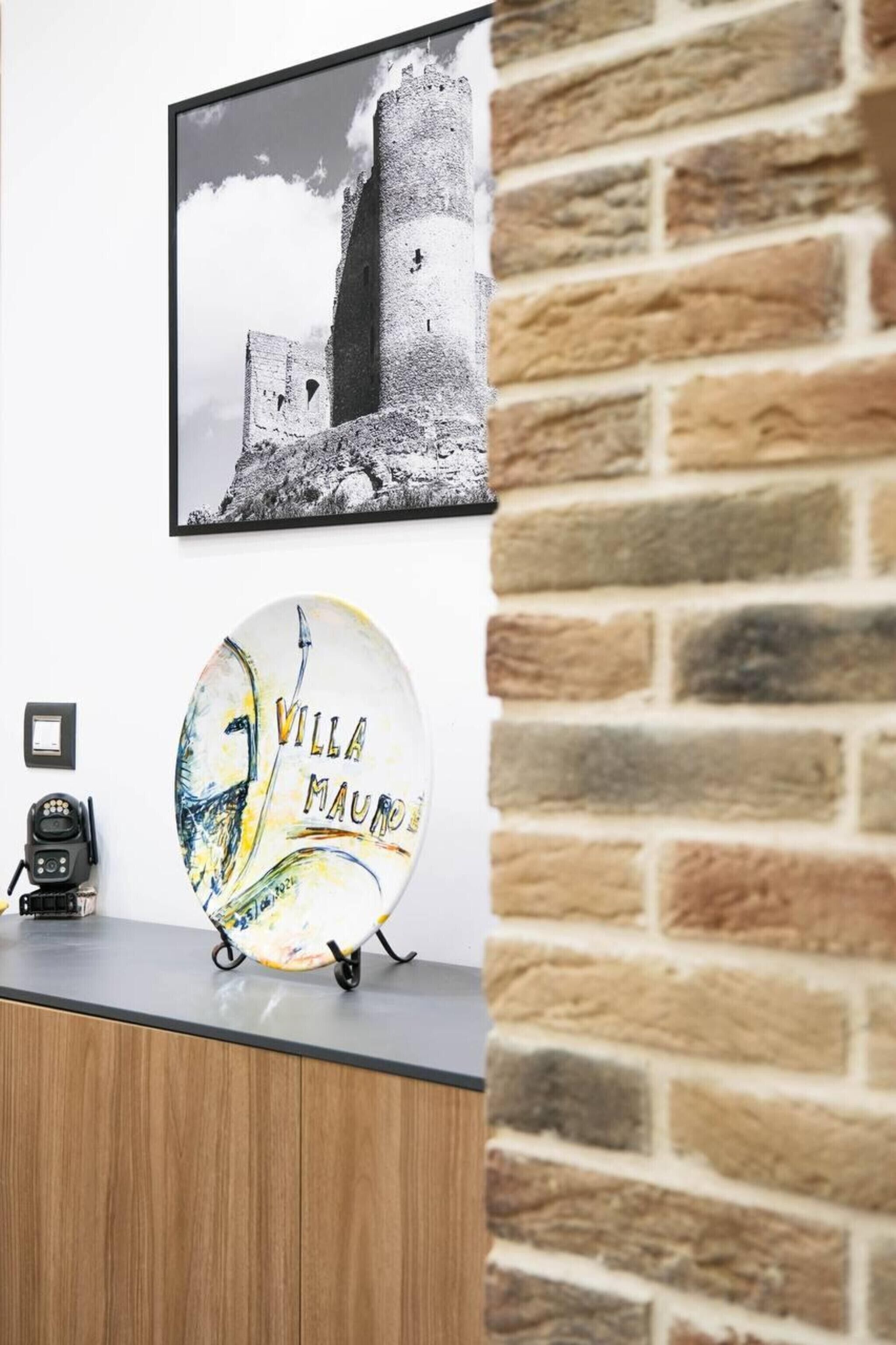 A wall-mounted picture of a castle, a round plate with 'VILLA MAURO' text, and a coffee machine on a counter.