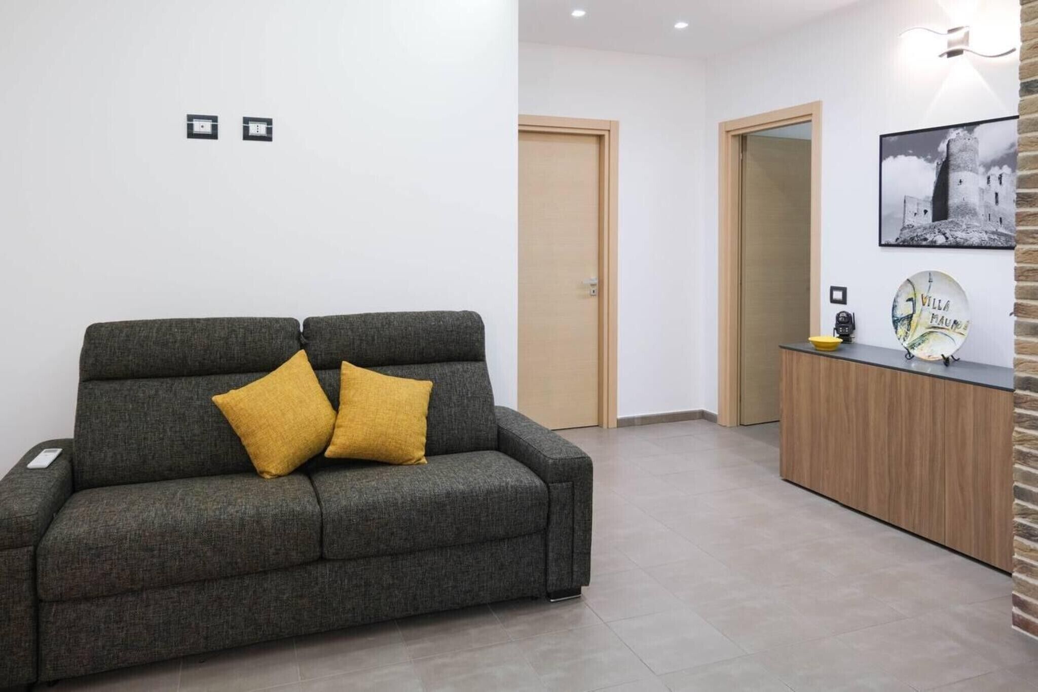 Een grijze bank met twee gele kussens, een houten kast, een ingelijst schilderij aan de muur, en een deur op de achtergrond.