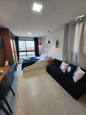 A modern hotel room with a bed, a black sofa, a dining area, and a view of the cityscape. - Villas do Porto - 209 by La Playa (Ipojuca)