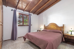 Een slaapkamer met een houten plafond, een bed met hoofdeinde, een raam met gordijnen, een nachtkastje met een lamp en een wandlamp.